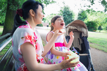 Portrait of young happy mother who sitting on bench outdoor and keeping on the laps beautiful daughter who is invalid and mother keeping cup of drink in hand