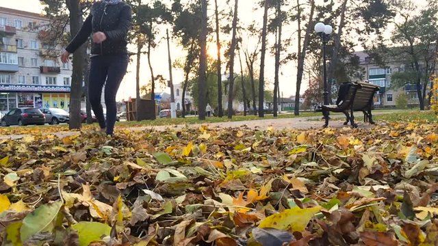 Woman walk and kick up yellow leaves by shoe tip, elegant slim legs in black shoes. Lady stroll at park in golden autumn time, play with leaf litter at lawn, throw up dry crunchy colourful leafage.