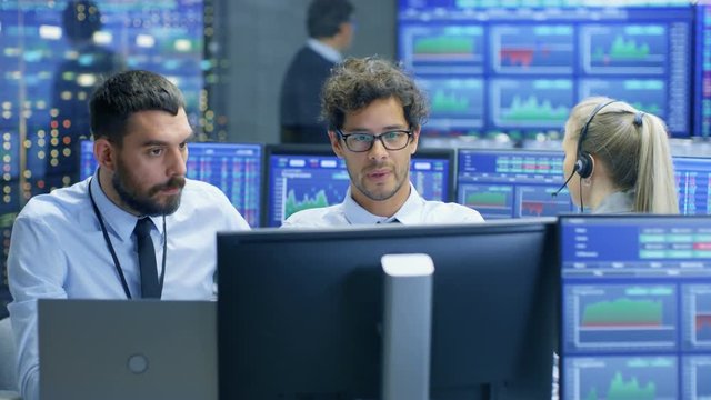 Professional Broker Consults Stock Exchange Trader at His Workstation. Multi-Ethnic Team at Stock Exchange Office is Busy Selling and Buying Stocks on the Market. Displays Show Relevant Data Numbers.
