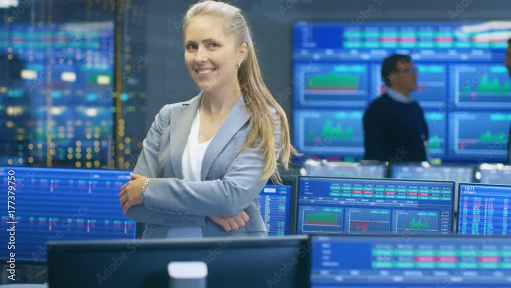 Poster successful female stock trader crosses arms and smiles at the camera. in the background busy stock e
