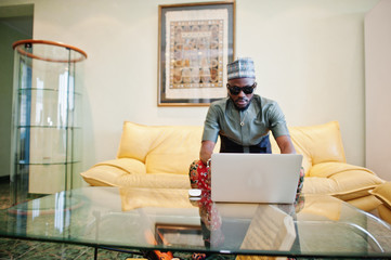 Rich african man sitting on sofa at his appartment and working on notebook.