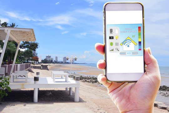 Hand holding smart phone for use application for control smart house system on long holiday background in the sea - Smart home technology of concept