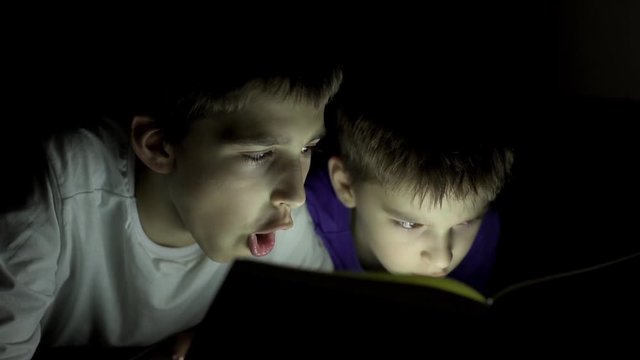 Two brothers reading a book hiding in the dark and using a torch light