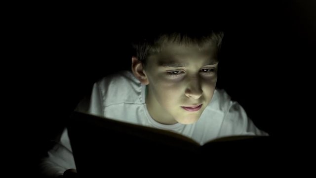 Young boy reading a book hiding in the dark and using a torch light