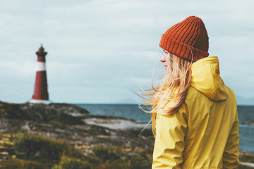 Woman sightseeing lighthouse sea landscape in Norway Travel Lifestyle concept scandinavian vacations outdoor. Blonde girl hair on wind wearing orange hat and yellow raincoat - obrazy, fototapety, plakaty