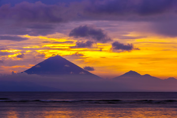 Naklejka na ściany i meble Colorful mountain silhouette against a bright orange sun, hidden behind blue clouds, the sea in the foreground. Gunung Agung mountain on sunset, Gili Trawangan, Indonesia.