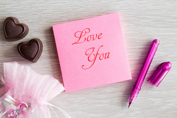 Reminder on the pink sticky note to a loved one with sweet hearts chocolate. Inscription Love you. Relationship concept with positive mood. Romantic atmosphere. Top view.
