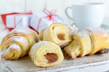 Homemade croissants with fruit jam, decorated with powdered sugar with a cup of morning tea on a white wooden background, scattered gift white and red boxes, tied with a twine.