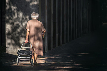 old woman walking alone, senior lady with cane and suitcases with wheels going into the darkness  