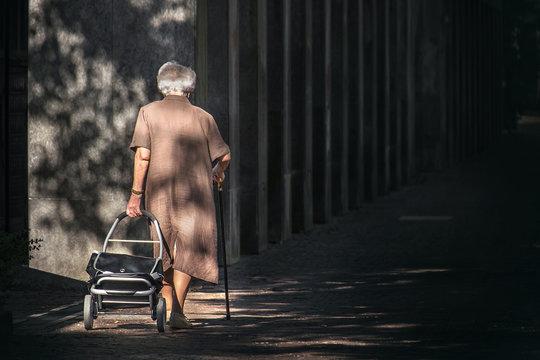 Old Woman Walking Alone, Senior Lady With Cane And Suitcases With Wheels Going Into The Darkness  