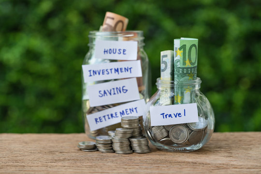 Selective focus on bottle jar with full of coins and banknote labeled as Travel and other jar in the background as saving for travel concept