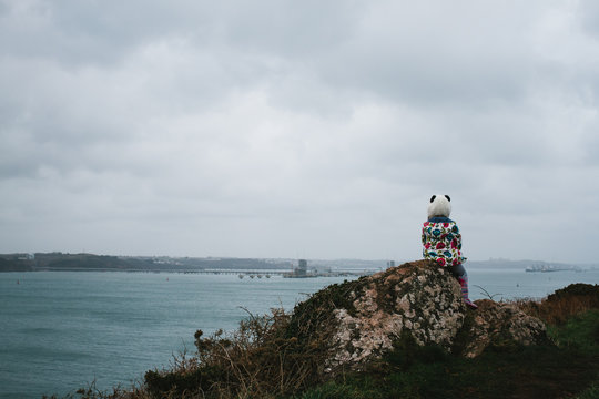 A little girl in a panda hat sitting on a rock looking at the sea.