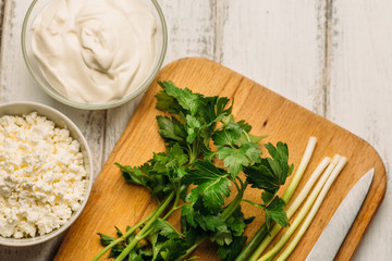 Top view of ingredients: sour cream, cheese curd and green herbs on cutting board on white rustic wooden table