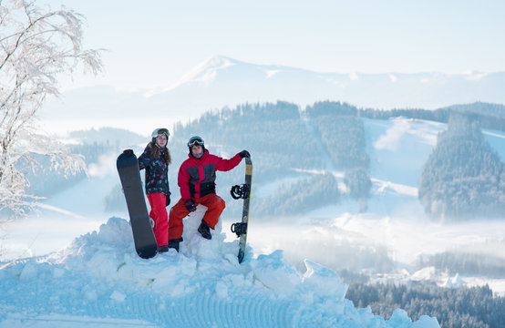 Pair of snowboarders on top of a mountain with a beautiful scenery of snow-capped mountains and forests of the Carpathians in a white haze. Guy is sitting on snowdrift, girl is standing next to him