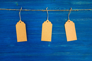 Three brown blank paper price tags or labels set hanging on a rope on the blue wooden background.