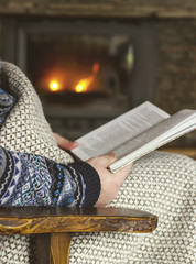 Obraz premium Man reading a book, sitting in a rocking chair with knit plaid in front of a fireplace