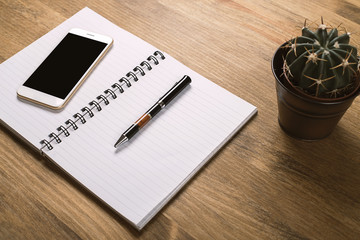 Smartphone with notebook and pen next to cactus on wooden table. Office.