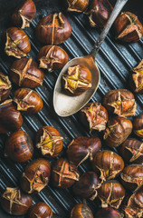Close-up of roasted chestnuts and metal vintage spoon over black cast iron grilling pan surface, top view, horizontal composition