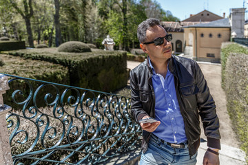 A man in leather jacket and black glasses looks around in a park while holding his mobile phone in his hand