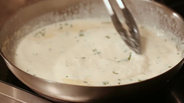 Stirring a Cream Sauce in a Frying Pan. a close up of someone stirring a cream sauce on a stove with tongs
