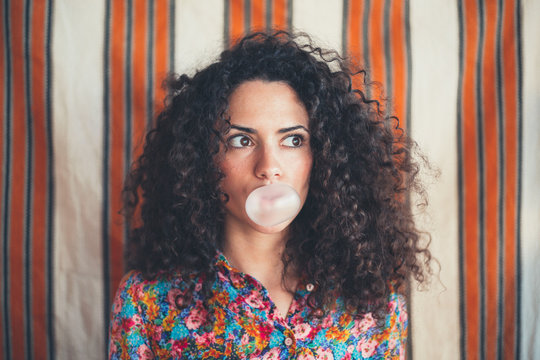 Girl with curly hair blowing a bubble with a pink bubble gum