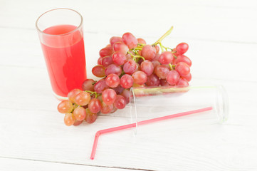Bunch of fresh ripe rose grapes near transparent and fragile glass full of juice beside empty glass with red tubule on old wooden rustic white planks