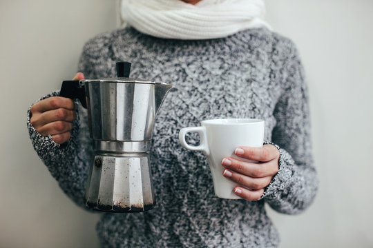 Woman with chunky knit sweater holding a hot cup of coffee and coffee pot. .
