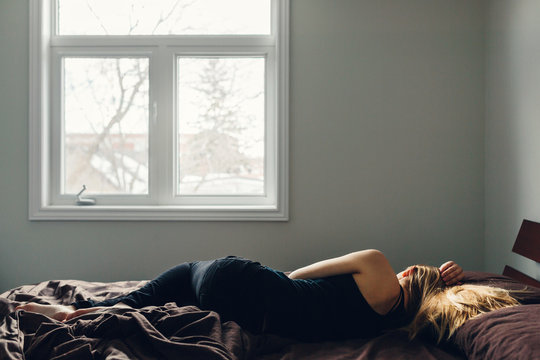 Blonde woman in a bedroom lying on a bed with rumpled sheets.