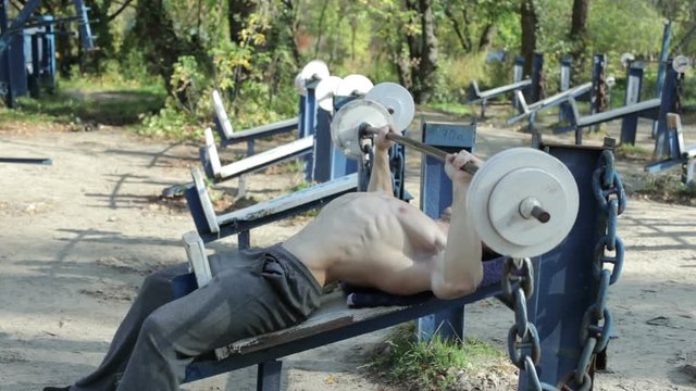 Bodybuilder trains with a barbell in a sports park outdoors.