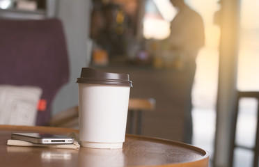 A white paper coffee cup putting on wooden table in coffee shop with a man ordering beverage as background.