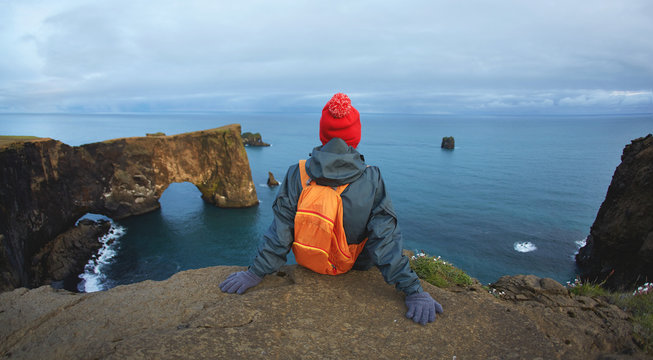 Girl in warm clothing, in red knitted hat and small orange backpack stands on the cliff on background of ocean, sky and Reynisfjara beach in Iceland. Popular tourist attraction. Unusual and gorgeous