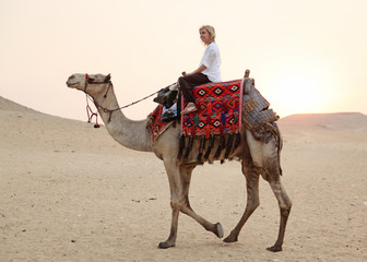 blonde Frau reitet auf einem Kamel durch die Wüste vor orangem Himmel