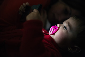 Baby using a mobile phone lying down on a bed and her mother on the background