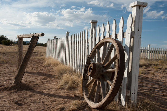 White wooden picket fence with wagon wheel resting against the fence in a rural setting.