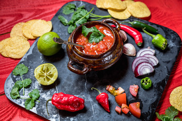 Homemade salsa with ingredients on black stone desk on red rustic background, tomato, pepper, onion, chili, habanero, coriander leaf. Mexican food concept