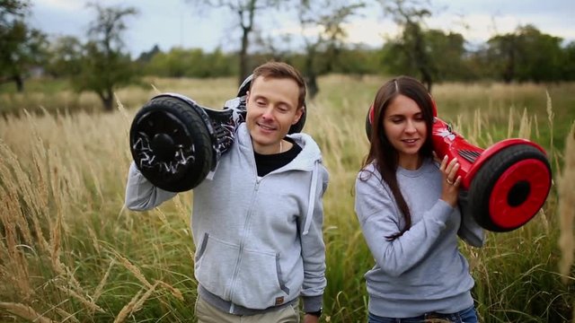 Young man and girl go into the field with hoverboards look at one another in urban clothing. Steadicam
