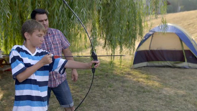 Adult camp leader watches as boy learning archery fires arrow from a professional, retail bow during an outdoor class at summer camp. Slow motion 4K recorded at 60fps.
