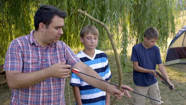 Adult camp leader teaches boys archery using a home-made bow during an outdoor class at summer camp. Boy in background uses more professional bow, 4K.