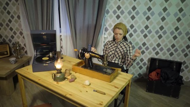 Retro seamstress girl winding threads, enjoys work, sews with old manual hand sewing machine. Woman working at home or workshop at night with kerosene lamp, listens music on gramophone phonograph