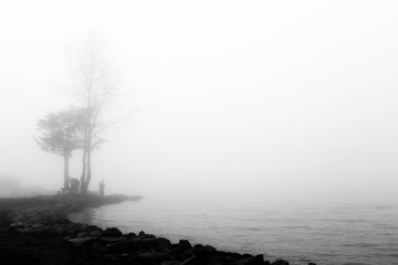 A lake hidden by fog, with rocks in the foreground and a fisherman and trees in the background
