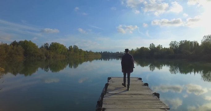 Giovane ragazzo con barba e cappello sta camminando su un molo di legno in riva a un lago 