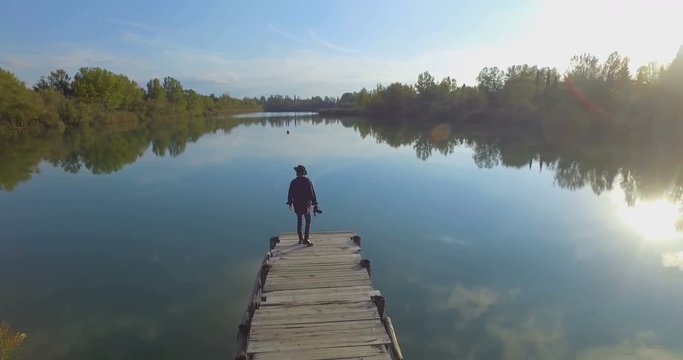 Giovane ragazzo con barba e cappello sta camminando su un molo di legno in riva a un lago 