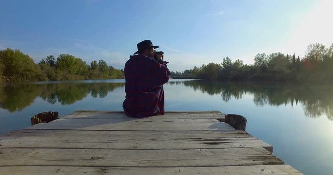 Giovane ragazzo con barba e cappello sta camminando su un molo di legno in riva a un lago 