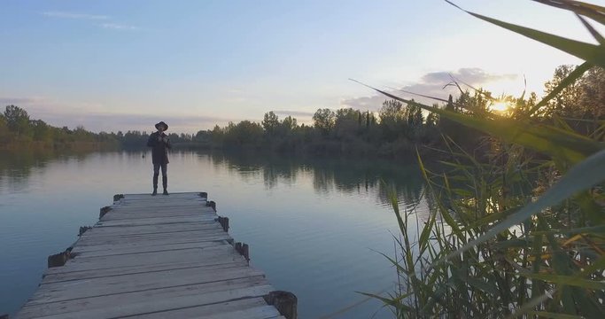 ragazzo con il cappello seduto su un molo di legno guarda tranquillo il lago blu al tramonto