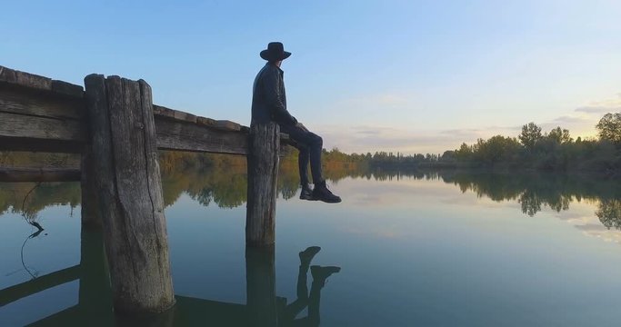 ragazzo con il cappello seduto su un molo di legno guarda tranquillo il lago blu al tramonto