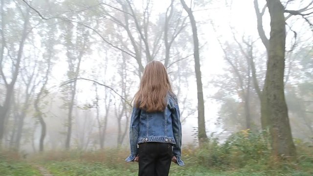 Little European girl with a long hair, blue jacket, black pants, sneakers and blue eyes. A frightened little child is standing in the foggy deserted forest. Loneliness. Steady cam fly around shot.