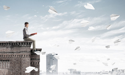 Handsome student guy on roof edge reading book and paper planes fly around