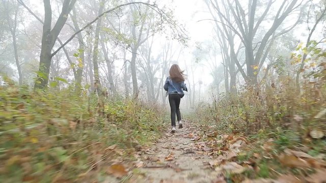 Little European girl with a long hair, blue jacket, black pants, sneakers and blue eyes. A frightened little child is running through the foggy deserted forest. Loneliness. Steady cam behind shot.