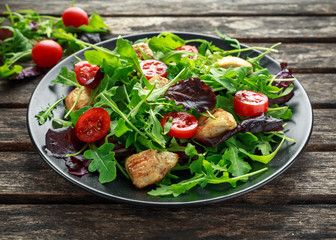 Fresh salad with chicken breast, arugula, nuts and tomatoes on black plate in a wooden table.