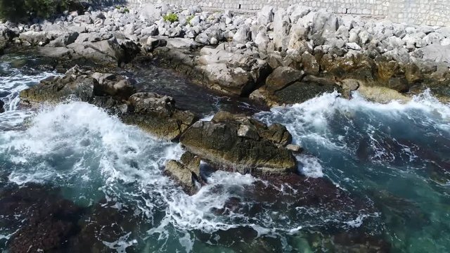 Low altitude aerial above rocky coastline wild sea hitting the rocks beautiful azure blue colored mediterranean ocean water and white foam by the power of water impact on stones amazing summer day 4k
